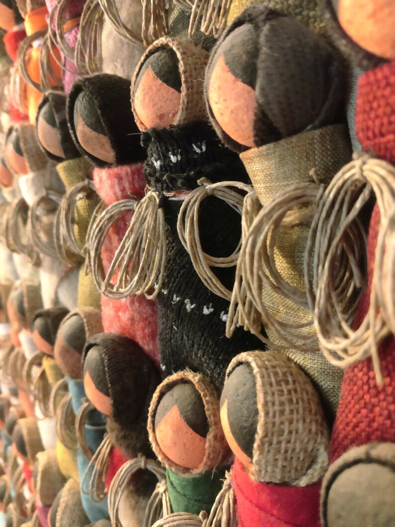 Pu&ograve; includere: Un'esposizione murale di bambole fatte a mano con diverse trame e colori di tessuto. Le bambole sono fatte di tessuto e hanno un design semplice con una testa rotonda e un corpo. Le bambole sono disposte in fila, con le teste delle bambole rivolte verso lo spettatore. Le bambole sono tutte di colori diversi, tra cui rosso, verde, blu e marrone.