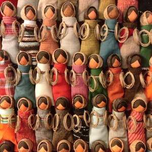 Pu&ograve; includere: Una collezione di bambole di stoffa colorate con teste di legno marrone e capelli di lana. Le bambole sono disposte in file e indossano tutte tessuti di diversi motivi e colori.