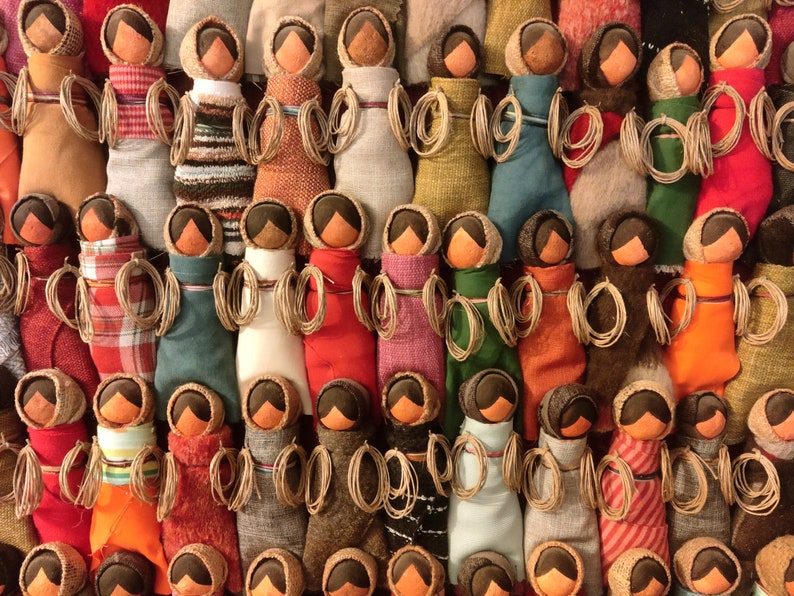 Pu&ograve; includere: Una collezione di bambole di stoffa colorate con teste di legno marrone e capelli di lana. Le bambole sono disposte in file e indossano tutte tessuti di diversi motivi e colori.