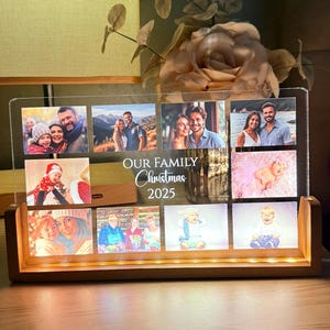 May include: A wooden photo display with a clear acrylic panel, lit by warm LED lights. It features multiple family photos and the text "OUR FAMILY Christmas 2025". The photos are arranged in a grid, creating a warm, inviting glow.