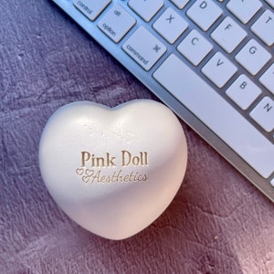 K&ouml;nnte beinhalten: Weiches, herzf&ouml;rmiges Spielzeug in Wei&szlig; mit dem Text "Pink Doll Aesthetics" in goldenen Buchstaben.
