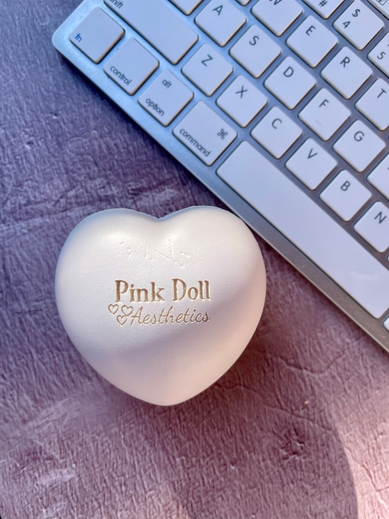 K&ouml;nnte beinhalten: Weiches, herzf&ouml;rmiges Spielzeug in Wei&szlig; mit dem Text "Pink Doll Aesthetics" in goldenen Buchstaben.