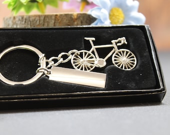 May include: Silver-colored bicycle keychain with a rectangular tag and a large key ring. The bicycle has detailed spokes and is presented in a black box with a velvet lining. The background includes greenery and flowers.
