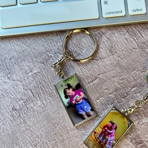 May include: Two silver keychains with rectangular photo inserts. The top keychain features a photo of a young girl wearing a pink shirt and blue shorts. The bottom keychain features a photo of two young girls wearing matching dresses.