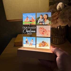 Puede incluir: Un expositor de fotos iluminado con seis fotos familiares. El expositor se apoya en una base de madera y presenta el texto "OUR FAMILY Christmas 2025". Las fotos muestran a miembros de la familia en la playa y con un beb&eacute;.