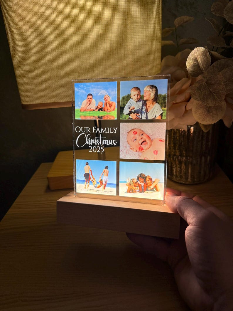 Puede incluir: Un expositor de fotos iluminado con seis fotos familiares. El expositor se apoya en una base de madera y presenta el texto "OUR FAMILY Christmas 2025". Las fotos muestran a miembros de la familia en la playa y con un beb&eacute;.