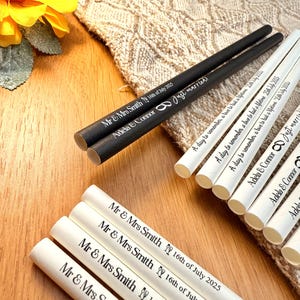 May include: A collection of personalized wedding straws. Some are black with "Mr & Mrs Smith 16th of July 2025" and "Adela & Connor Just married!" printed in white. Others are white with similar text. A yellow flower is in the background.