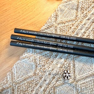 May include: Three black pencils with the inscription "A day to remember, a love to last a lifetime. 25th July 2025" in white lettering. The pencils rest on a textured, beige and white patterned surface.