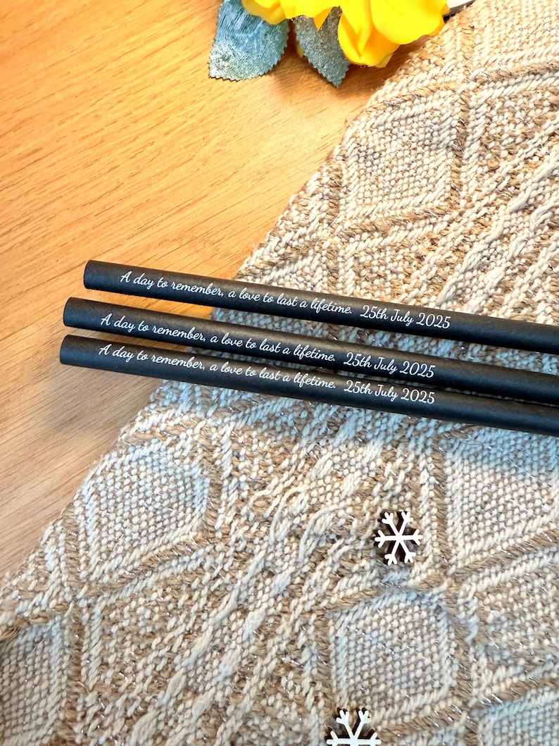 May include: Three black pencils with the inscription "A day to remember, a love to last a lifetime. 25th July 2025" in white lettering. The pencils rest on a textured, beige and white patterned surface.