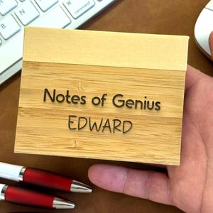 May include: A bamboo notepad with the text "Notes of Genius EDWARD" engraved on the front. The notepad is rectangular with a light tan top and a natural wood grain base. Three red and silver pens are visible below the notepad.