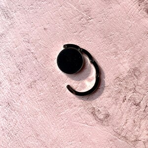 May include: A silver-toned metal bag clip with a black circular button. The clip is designed to be used for securing bags or pouches.