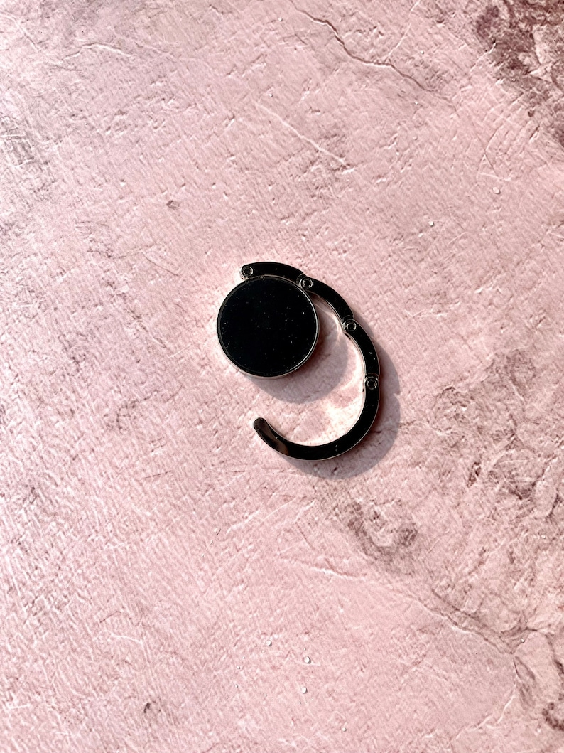 May include: A silver-toned metal bag clip with a black circular button. The clip is designed to be used for securing bags or pouches.