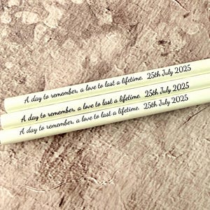 May include: Three white pencils with the inscription "A day to remember, a love to last a lifetime. 25th July 2025" in black script. The pencils are arranged diagonally against a textured, neutral-toned background.