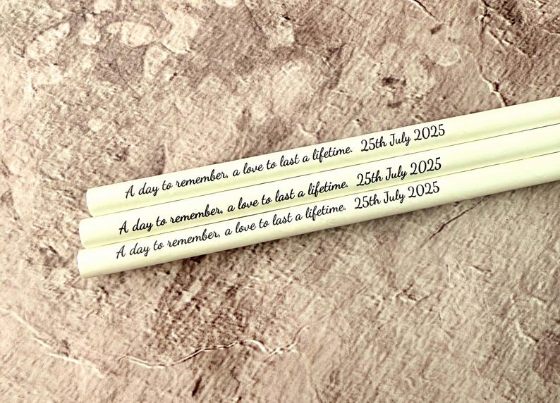 May include: Three white pencils with the inscription "A day to remember, a love to last a lifetime. 25th July 2025" in black script. The pencils are arranged diagonally against a textured, neutral-toned background.