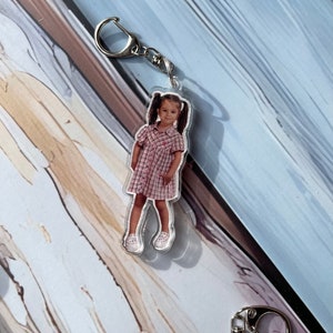 May include: A clear acrylic keychain featuring a photo of a young girl wearing a pink and white plaid dress and white shoes. The keychain has a silver metal ring.