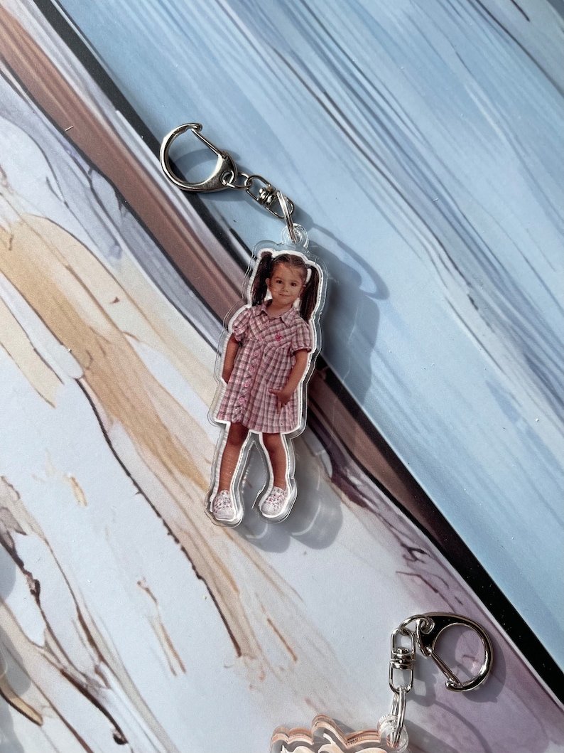 May include: A clear acrylic keychain featuring a photo of a young girl wearing a pink and white plaid dress and white shoes. The keychain has a silver metal ring.