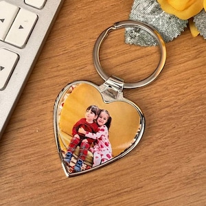 May include: Silver heart-shaped keyring featuring a photograph of two children embracing. The photo shows the children in red and pink clothing. The background is yellow. The keyring has a large, circular ring for keys.