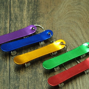 May include: Five colourful miniature skateboard keychains, each with a different colour: purple, blue, gold, green, and red. The keychains are arranged in a row on a wooden surface.
