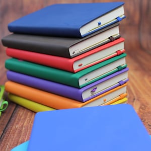 May include: A stack of nine colourful notebooks with different coloured covers, including blue, grey, red, green, purple, orange, and yellow. The notebooks are stacked on a wooden surface.