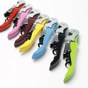 May include: A set of seven colourful wine bottle openers. The openers are made of metal with a plastic handle. The handles are red, pink, maroon, yellow, blue, black, and green.