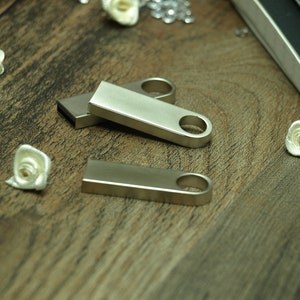 May include: Three silver metal USB flash drives with a loop on the end. The flash drives are on a wooden surface.