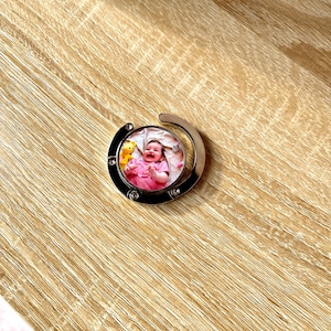 May include: A silver-toned retractable keychain with a round photo insert featuring a baby in a pink dress. The photo insert is surrounded by a silver metal frame.