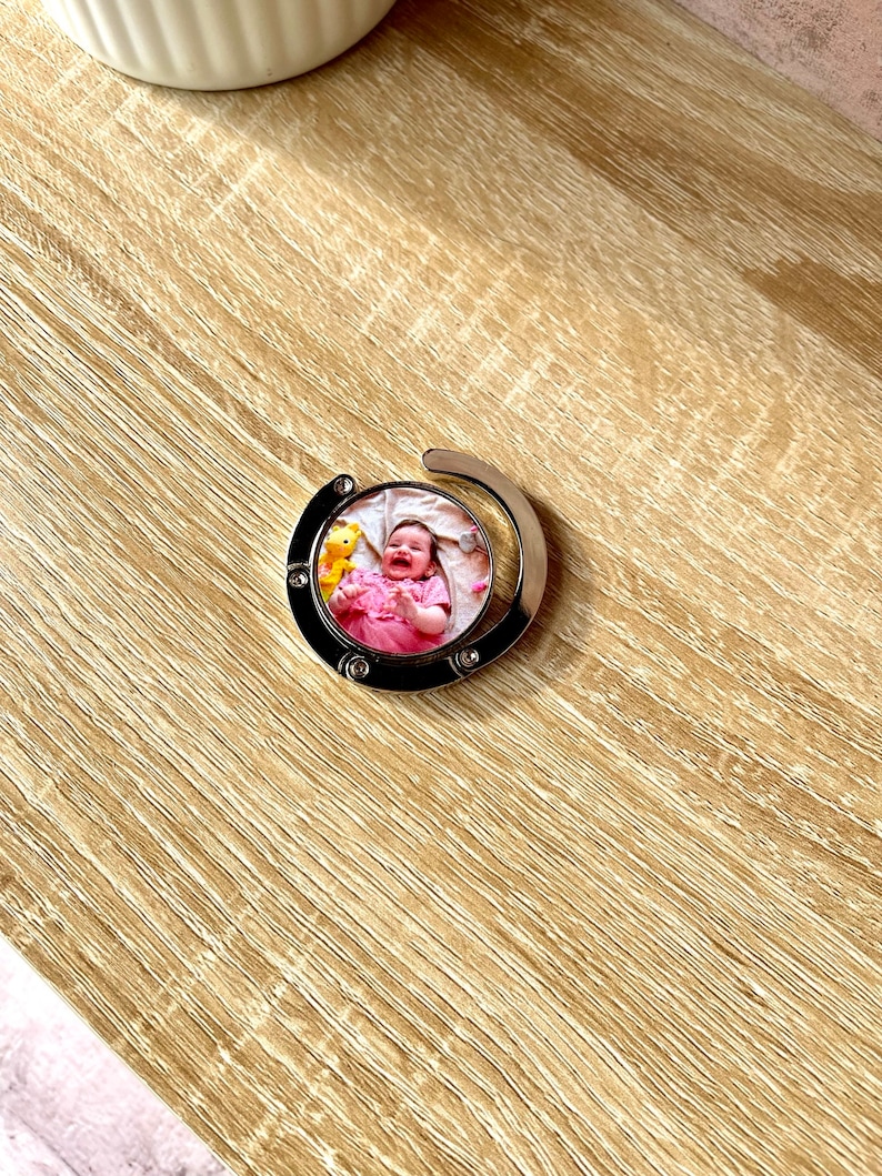 May include: A silver-toned retractable keychain with a round photo insert featuring a baby in a pink dress. The photo insert is surrounded by a silver metal frame.