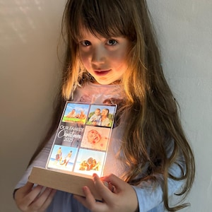 Puede incluir: Una pantalla fotogr&aacute;fica acr&iacute;lica iluminada con fotos familiares y el texto "OUR FAMILY Christmas 2025". La pantalla tiene una base de madera y la sostiene una persona. Las fotos son de una familia.