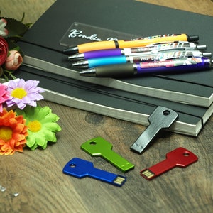 May include: Four colorful key-shaped USB drives on a wooden surface. The drives are blue, green, red, and silver. A black notebook with a silver pen holder and several pens is in the background.
