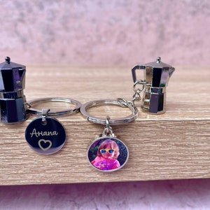 May include: Two silver metal coffee pot keyrings with a silver keyring. One keychain has a round silver charm with the name "Ariana" engraved on it and a small heart. The other keychain has a round silver charm with a photo of a young girl wearing sunglasses.