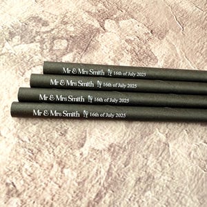 May include: Four dark gray paper straws are stacked, each printed with "Mr & Mrs Smith 16th of July 2025" in white. The straws are on a textured, light brown surface.