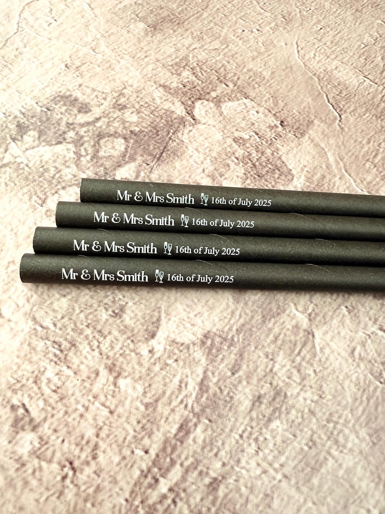 May include: Four dark gray paper straws are stacked, each printed with "Mr & Mrs Smith 16th of July 2025" in white. The straws are on a textured, light brown surface.