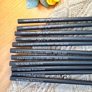 May include: A collection of black pencils with white text, featuring wedding-themed phrases like "Just married!" and dates. The pencils are arranged on a textured surface, with a yellow flower and decorative elements in the background.