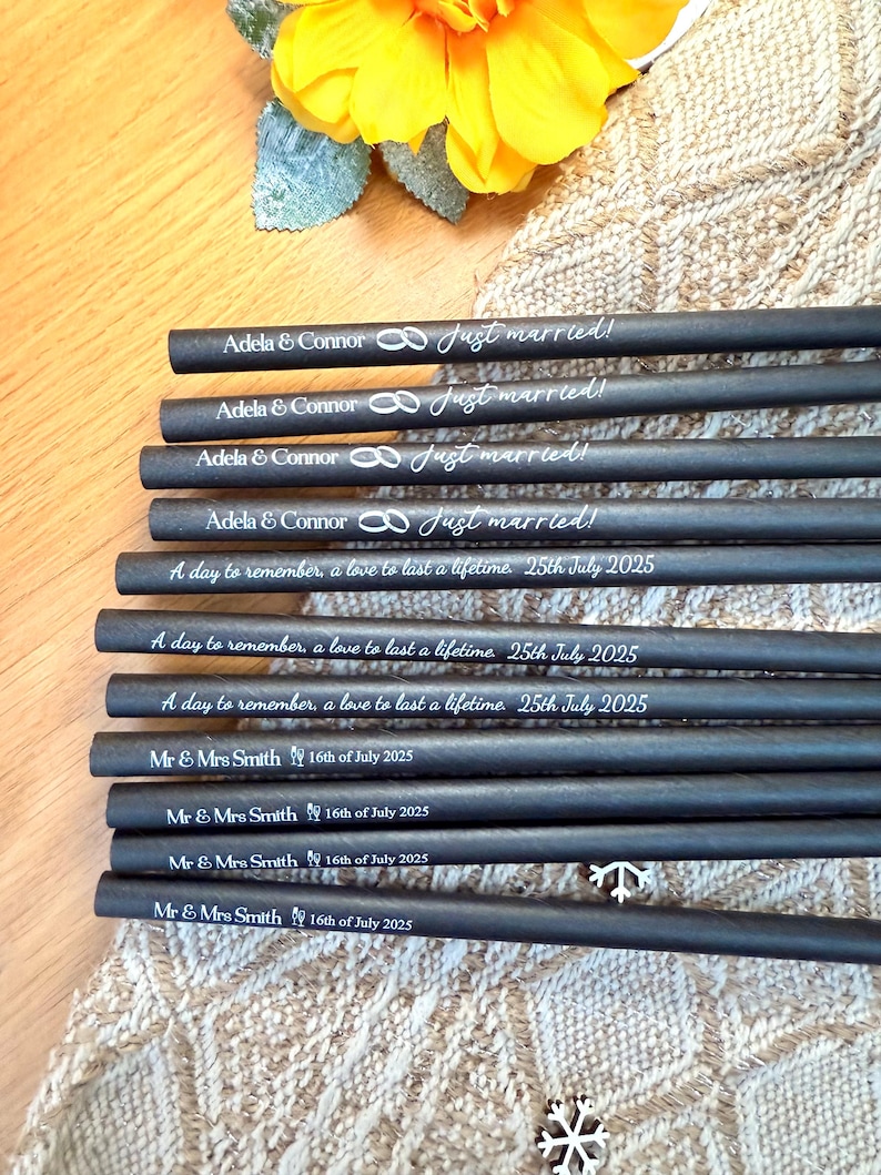 May include: A collection of black pencils with white text, featuring wedding-themed phrases like "Just married!" and dates. The pencils are arranged on a textured surface, with a yellow flower and decorative elements in the background.