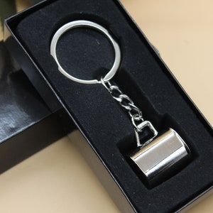 May include: A silver keyring with a small, cylindrical charm. The keyring is in a black box with a black velvet lining.