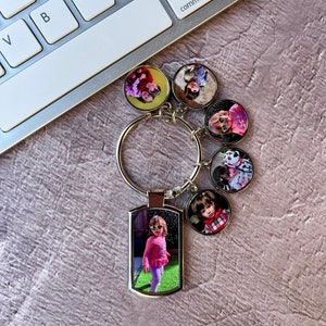 May include: A silver keyring with a rectangular charm featuring a photo of a young girl wearing sunglasses and a pink shirt. The keyring also has five round charms with photos of children.