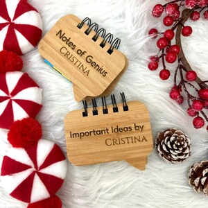 May include: Two small, bamboo-style notebooks with black spiral binding. One reads "Notes of Genius CRISTINA" and the other "Important Ideas by CRISTINA". The notebooks are on a white, fluffy surface, with red and white peppermint candies and a sprig of red berries.