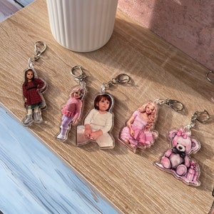 May include: Five keychains with different designs. The keychains feature a girl in a red dress, a girl in a pink dress, a girl in a white shirt, a girl in a pink dress, and a pink teddy bear holding a pink gift box.