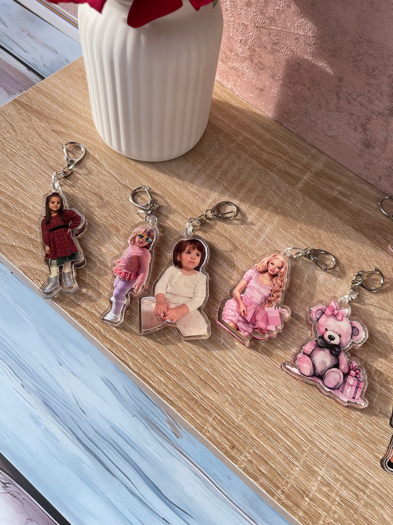 May include: Five keychains with different designs. The keychains feature a girl in a red dress, a girl in a pink dress, a girl in a white shirt, a girl in a pink dress, and a pink teddy bear holding a pink gift box.