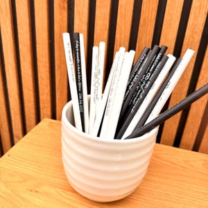 May include: A white ceramic cup filled with black and white pencils. The pencils have text printed on them, including phrases like "Just married!" and "A day to remember, a love to last a lifetime." The cup sits on a wooden surface.