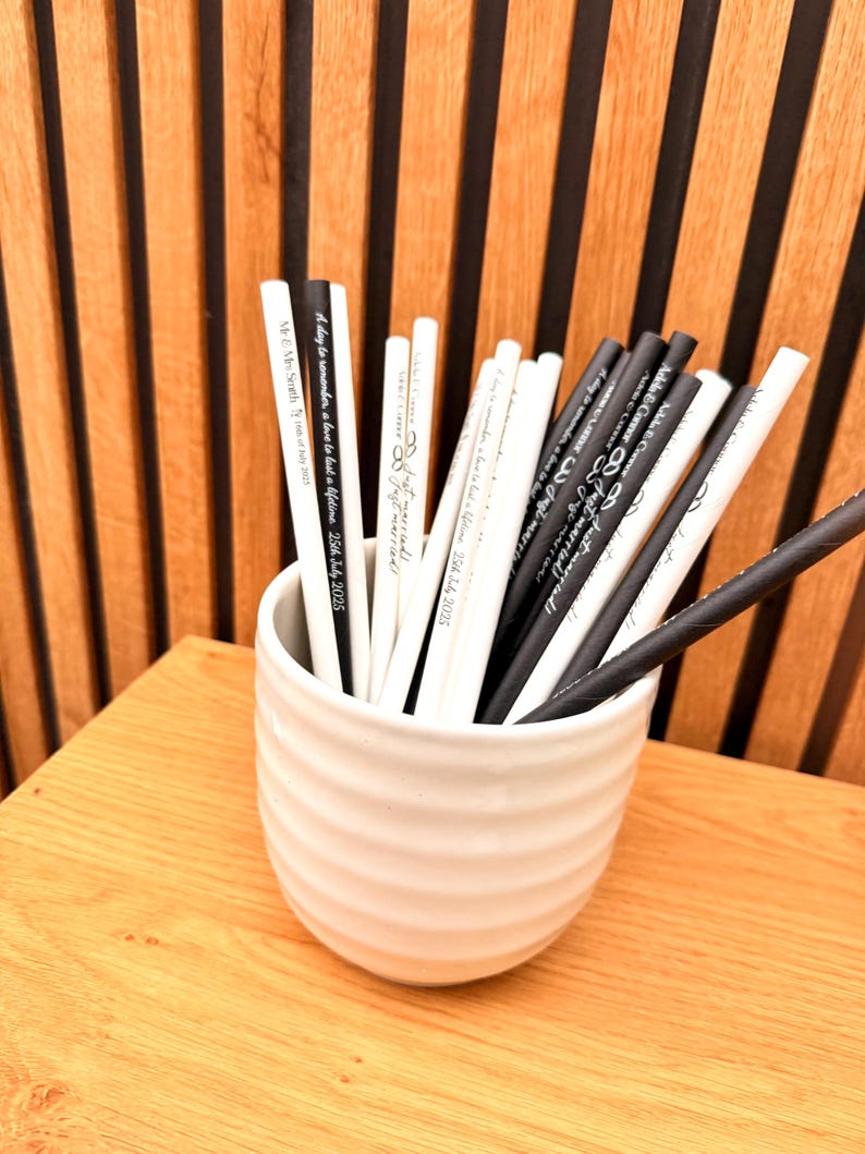 May include: A white ceramic cup filled with black and white pencils. The pencils have text printed on them, including phrases like "Just married!" and "A day to remember, a love to last a lifetime." The cup sits on a wooden surface.