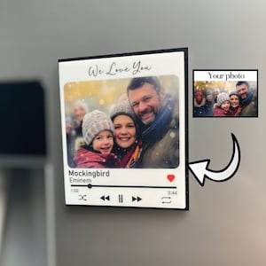 May include: A white rectangular plaque featuring a family photograph and the words "We Love You." Below the photo is a music player interface displaying the song "Mockingbird" by Eminem. A small photo is in the upper right.