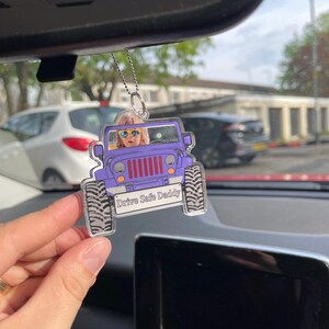 May include: A clear acrylic car air freshener in the shape of a purple Jeep. It features a photo of a person wearing sunglasses and the text "Drive Safe Daddy". The air freshener hangs from a silver chain.