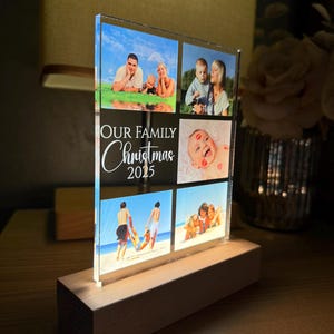 Puede incluir: Un expositor de fotos de acr&iacute;lico transparente con seis fotos familiares, montado sobre una base de madera. El texto "OUR FAMILY Christmas 2025" est&aacute; impreso en la parte delantera. Las fotos muestran a miembros de la familia en varios entornos.