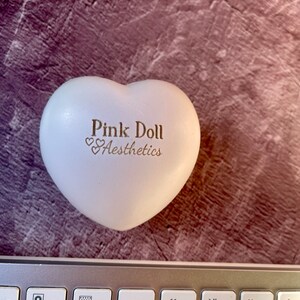 K&ouml;nnte beinhalten: Wei&szlig;er herzf&ouml;rmiger Stressball mit dem Text "Pink Doll Aesthetics" in goldenen Buchstaben.
