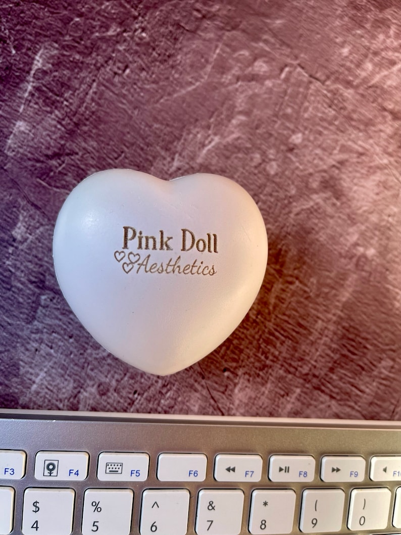 K&ouml;nnte beinhalten: Wei&szlig;er herzf&ouml;rmiger Stressball mit dem Text "Pink Doll Aesthetics" in goldenen Buchstaben.