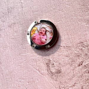 May include: A silver metal keychain with a round photo insert featuring a baby girl smiling and holding a stuffed animal. The photo is surrounded by a white border.