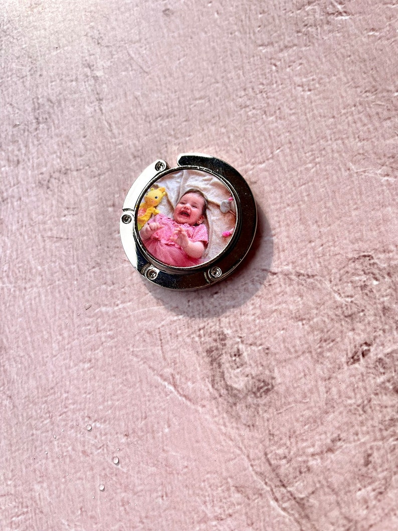 May include: A silver metal keychain with a round photo insert featuring a baby girl smiling and holding a stuffed animal. The photo is surrounded by a white border.