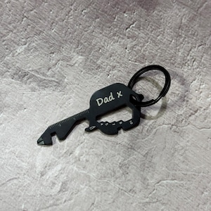 May include: Black metal key-shaped multi-tool keychain with a bottle opener, ruler, and screwdriver. The keychain has a ring for attaching to keys and is engraved with the text "Dad x".