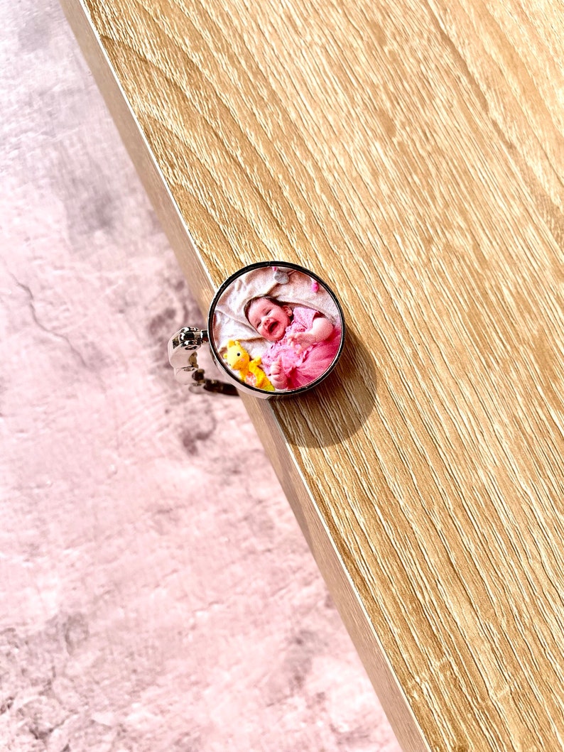 May include: A silver phone ring holder with a round photo insert featuring a baby in a pink outfit holding a yellow stuffed animal.